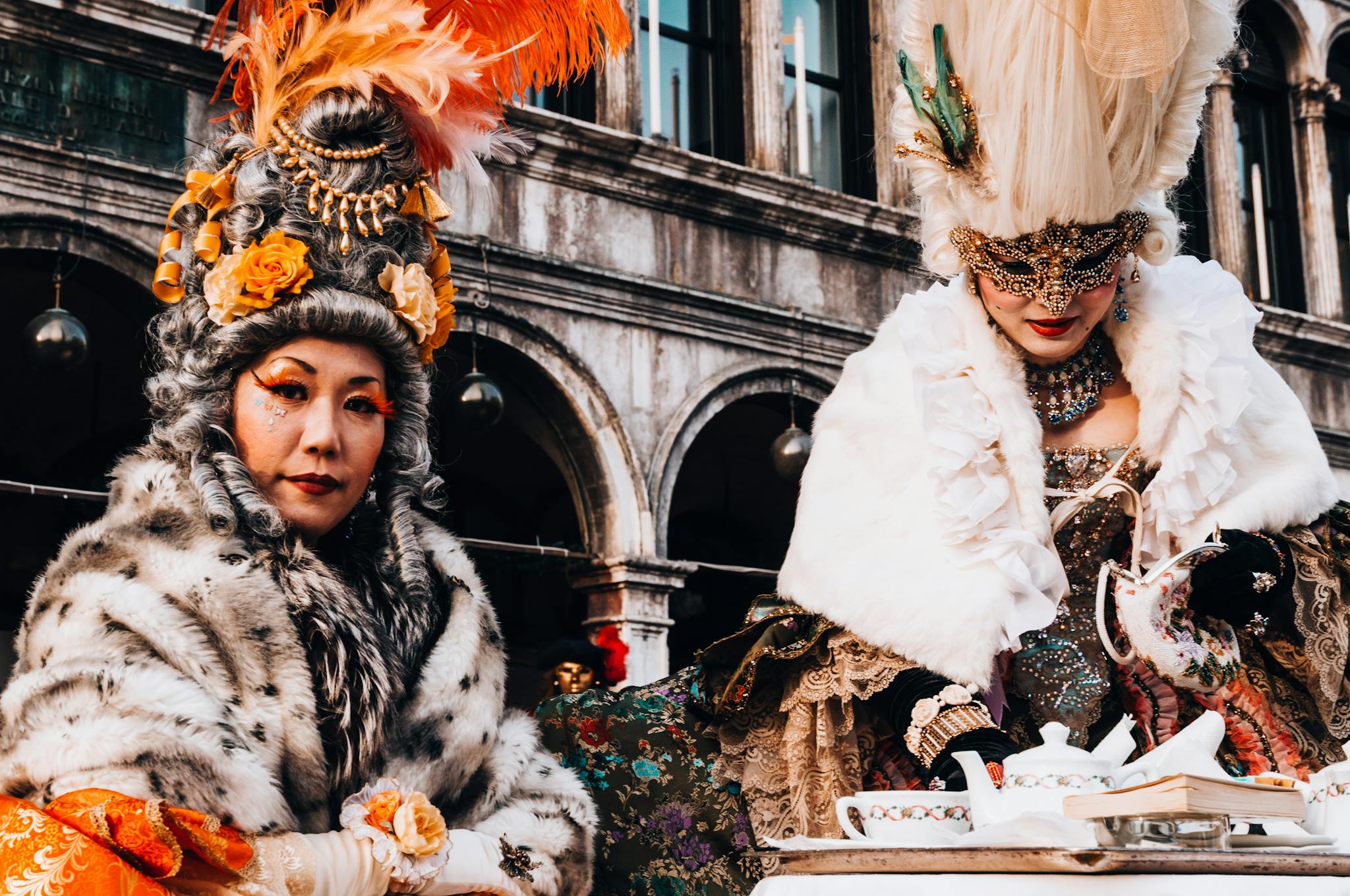 Two women in elaborate Venetian costumes enjoying a tea party during a masquerade festival.